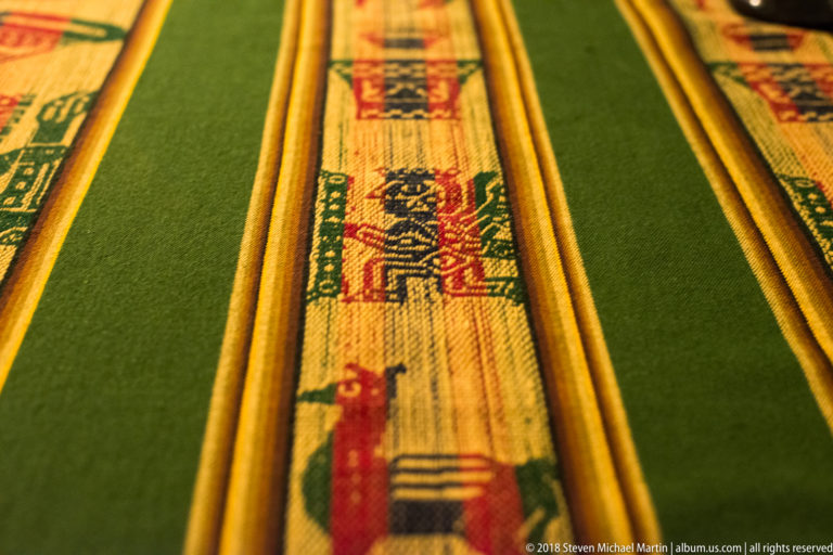 Close-up of green woven textile with narrow multicolored patterned bands and small stylized figures.