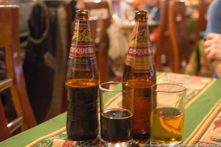 Two Cusqueña beer bottles with small glasses of dark and pale beer on a woven table runner.