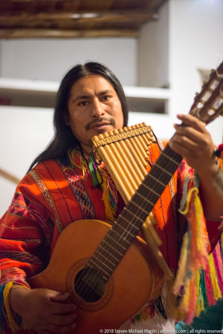 Andean musician in bright woven clothing playing panpipes while holding a guitar.