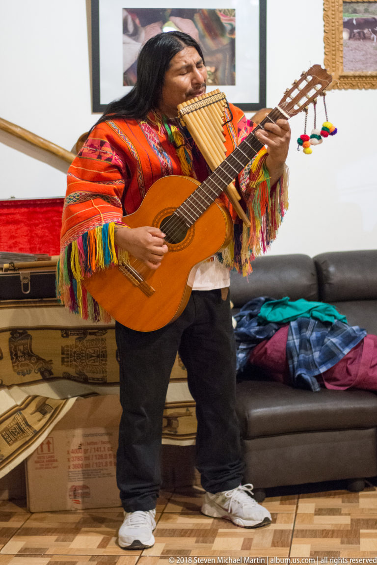 Male musician in a red woven poncho playing guitar and panpipes indoors.