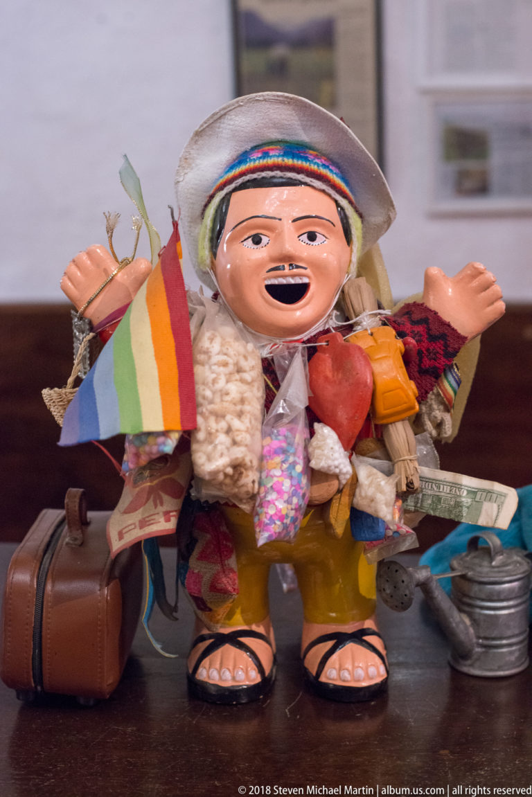 Colorful souvenir figurine wearing a broad hat and carrying miniature luggage, goods, and a rainbow-striped flag.