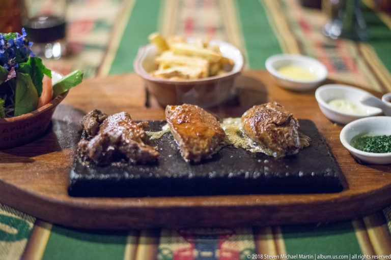 Three pieces of browned meat on a black hot stone with fries, salad, and small bowls of sauce.