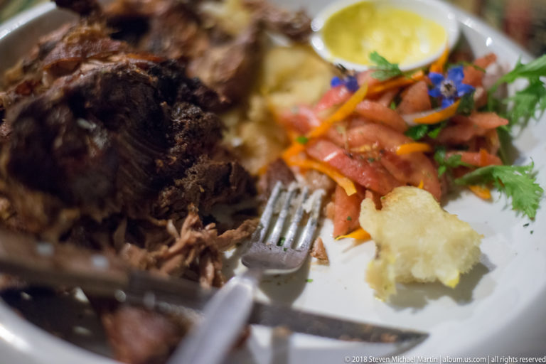 Roasted meat with tomato salad, potato, and yellow sauce on a white plate, with a fork cutting into it.