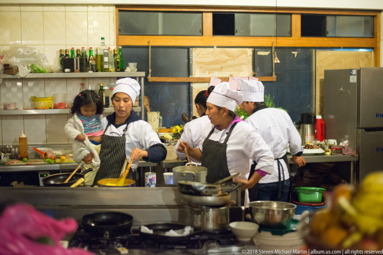 Several cooks preparing food in an open kitchen with pans, bowls, shelves, and stainless-steel equipment.
