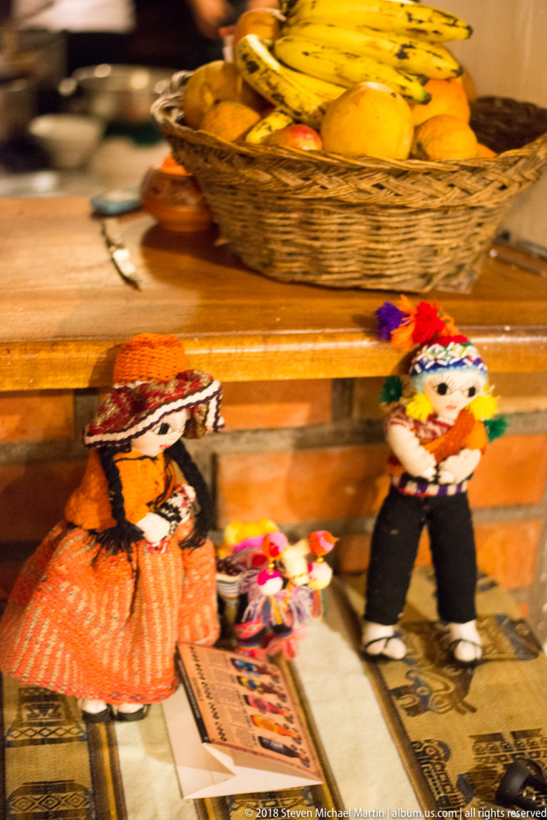 Colorful handmade textile dolls displayed on a patterned surface below a basket of yellow fruit.