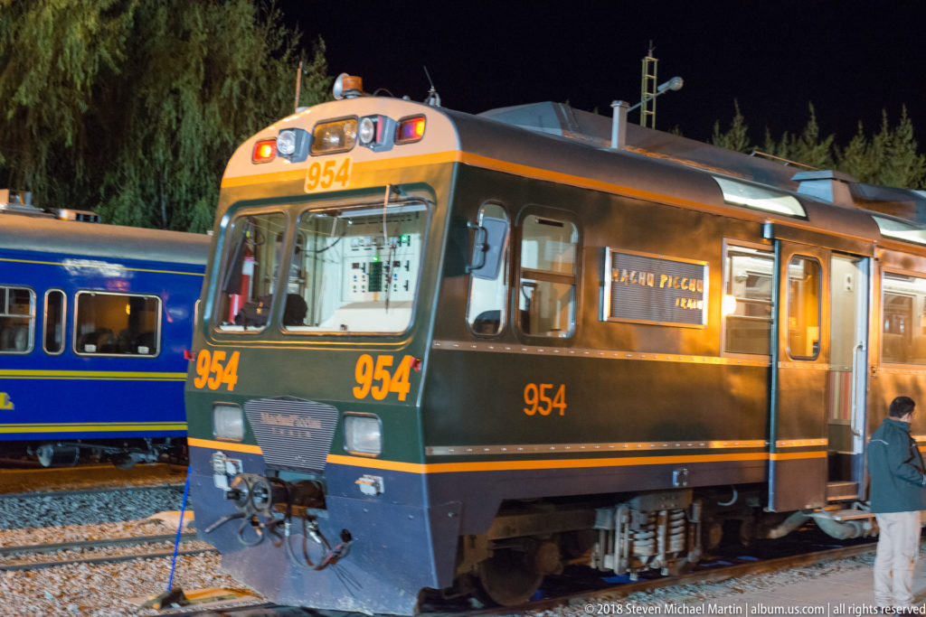 Dark green-and-orange rail vehicle number 954 beside a blue passenger coach at night.