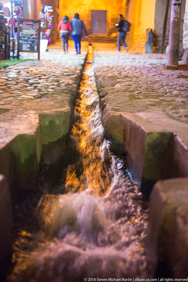 Fast-flowing water in a narrow stone channel crossing a cobbled street, with pedestrians beyond at night.