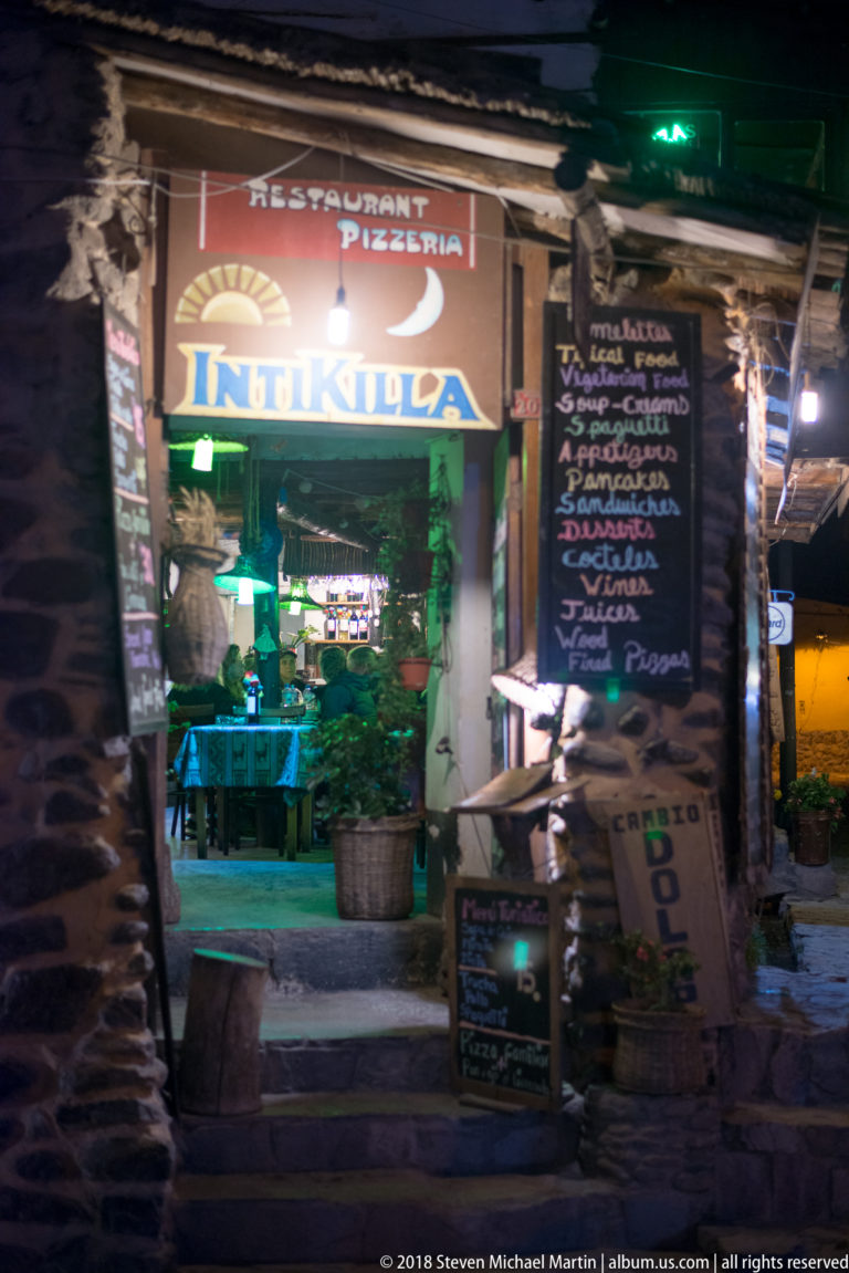 Nighttime restaurant or pizzeria entrance with illuminated Italian-themed sign, chalkboard menus, and tables inside.