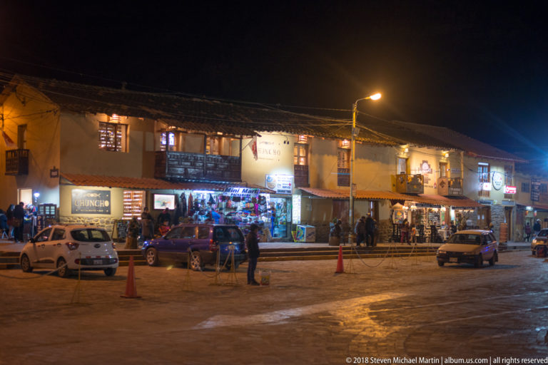 Night streetscape with two-story shops, parked cars, pedestrians, signs, and balconies around an open paved area.