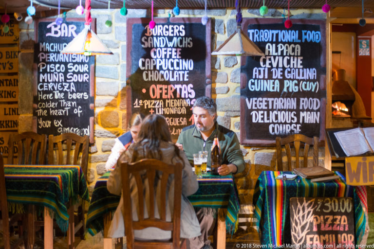 Two diners at a striped table beneath chalkboard menus listing coffee, alpaca, ceviche, guinea pig, and other dishes.
