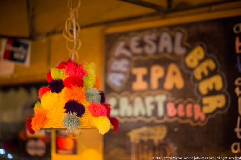 Conical hanging ornament made from bright multicolored yarn pom-poms, with a blurred beer sign behind.