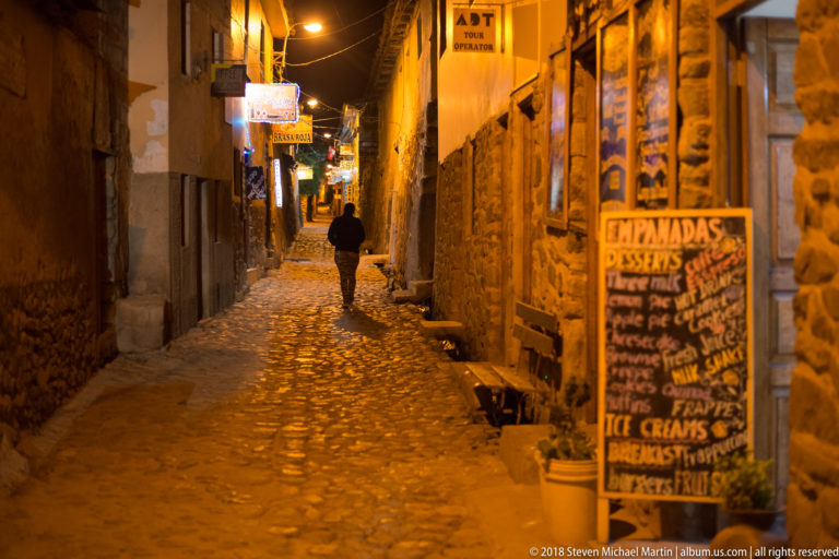 Narrow cobbled street at night with restaurant signs, stone walls, menu boards, and a lone pedestrian.