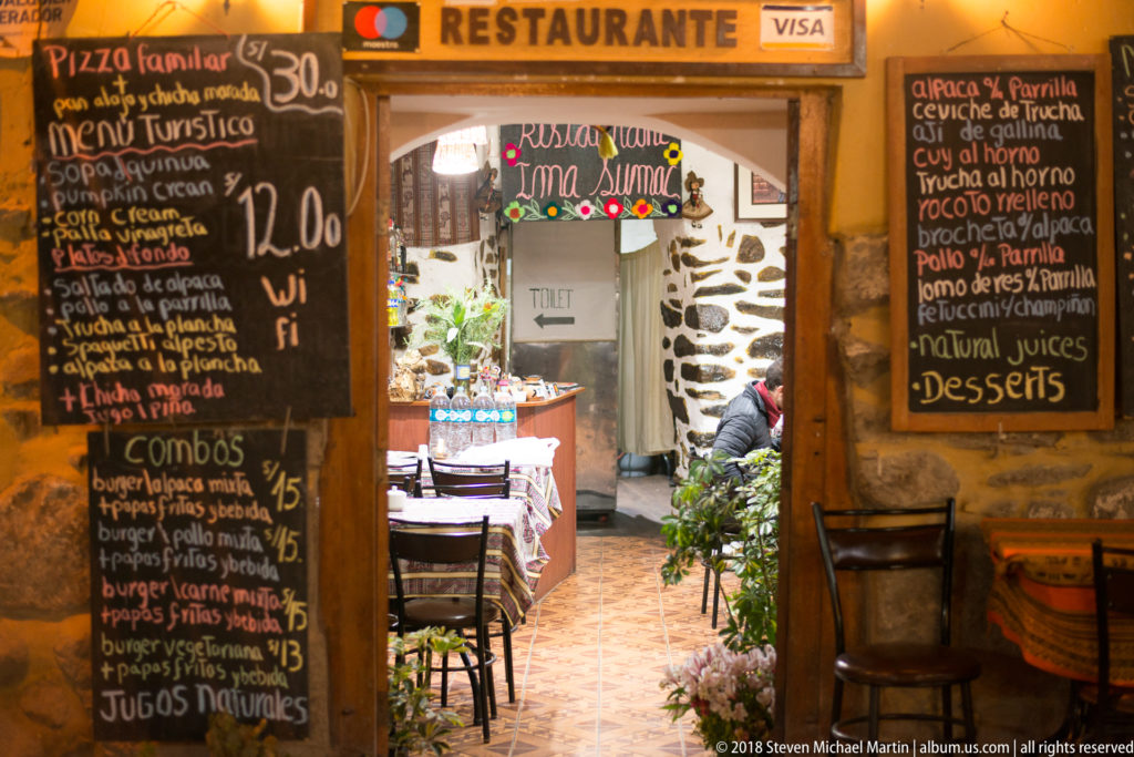 Restaurant entrance flanked by large handwritten menu boards, with tables visible inside under warm lighting.