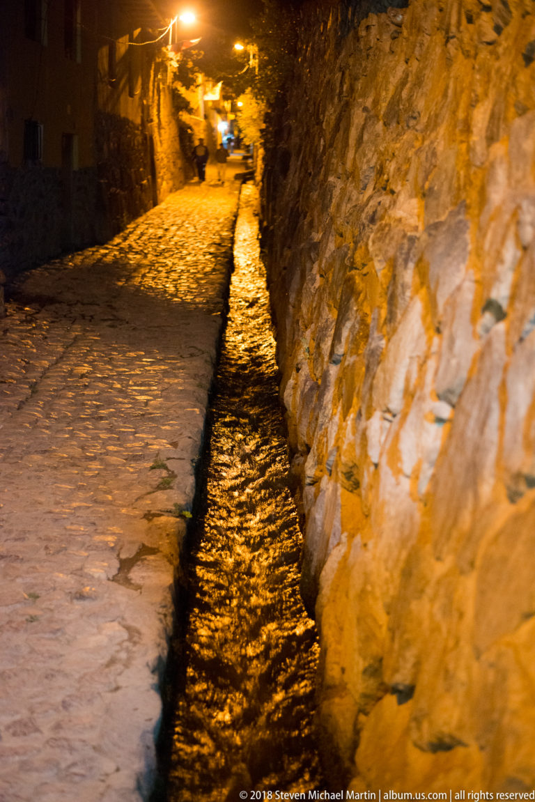 Stone-lined canal flowing beside a cobbled lane and high masonry wall under amber streetlights at night.