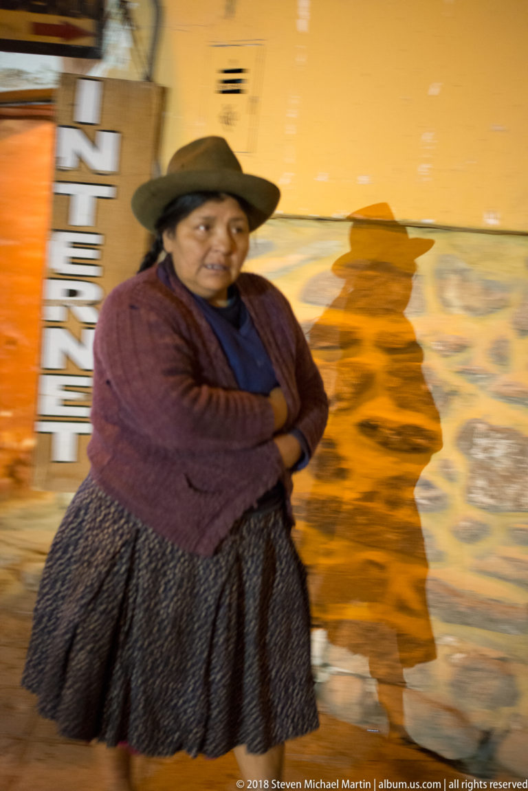 Woman in broad dark hat, burgundy cardigan, blue top, and patterned skirt standing against a painted wall.