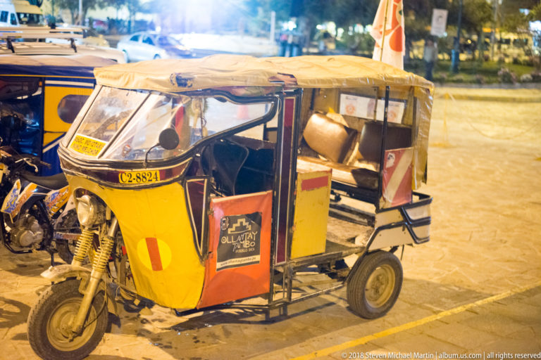 Yellow three-wheeled mototaxi with canopy and Peruvian flag decal parked on a street at night.