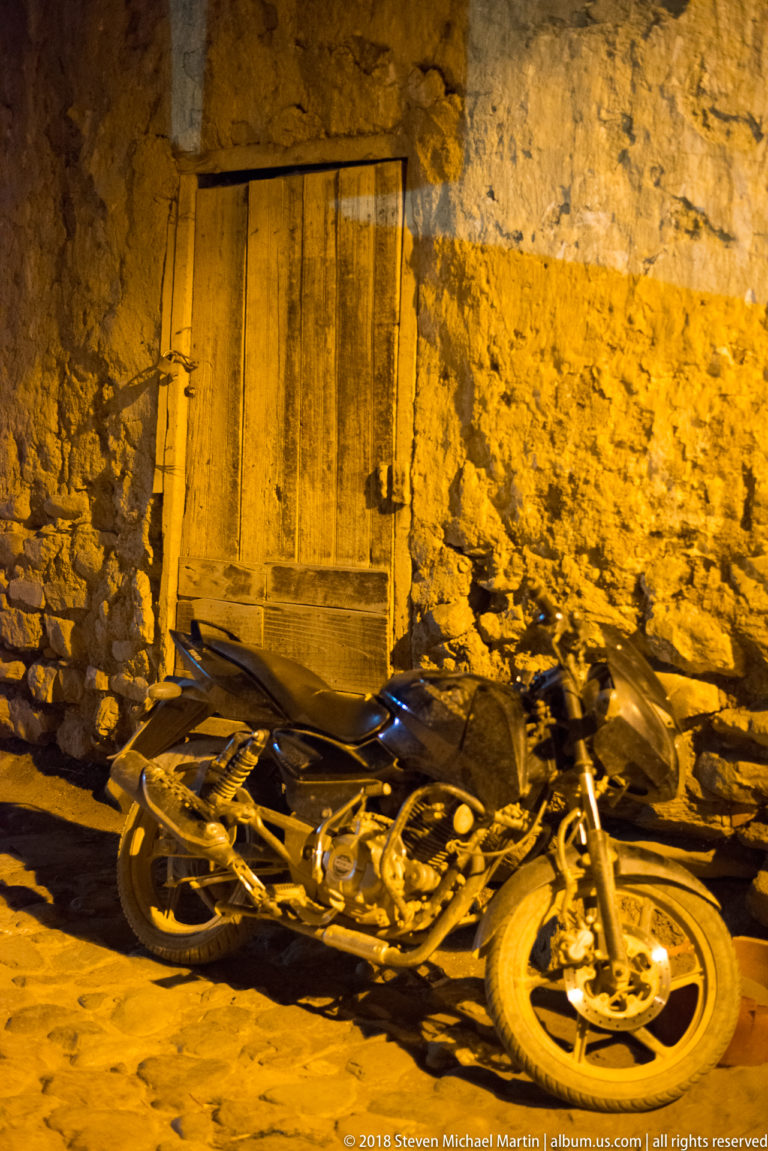 Motorcycle parked beside a rough stone wall and closed wooden door under warm nighttime lighting.