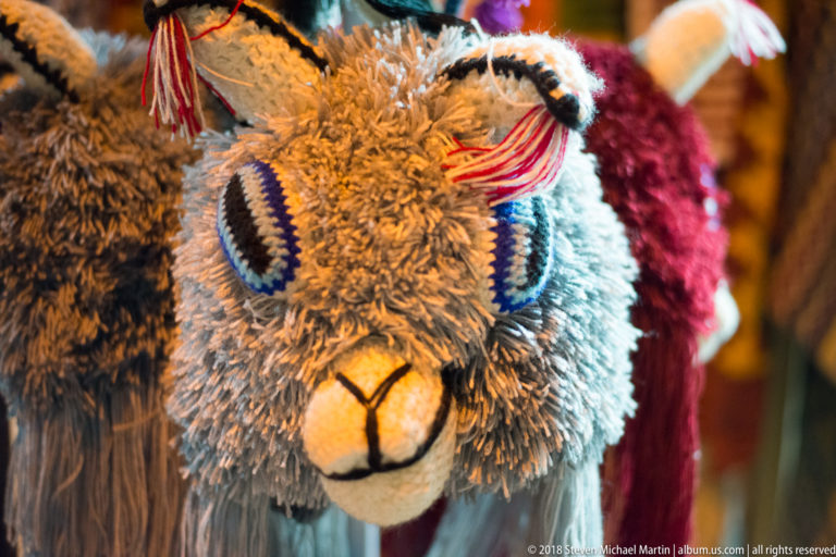 Close-up of a shaggy alpaca or llama souvenir with blue woven eyes, pale muzzle, and red tassel.