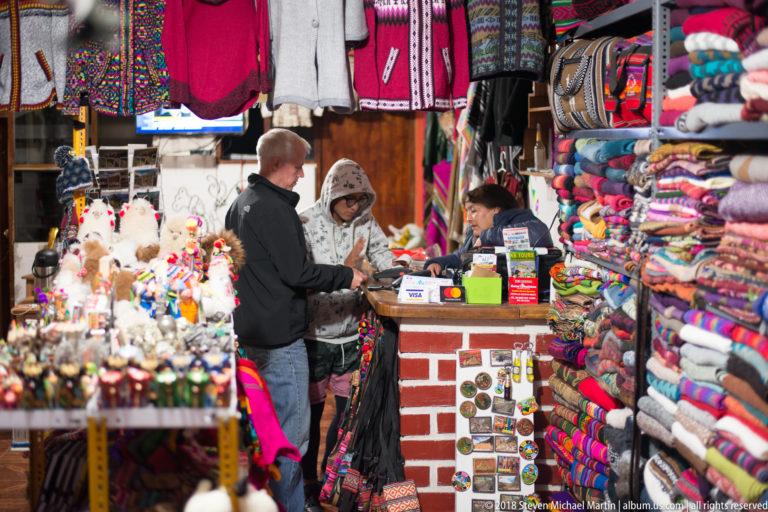 Customer at a souvenir shop counter surrounded by folded textiles, hats, bags, plush animals, and handicrafts.
