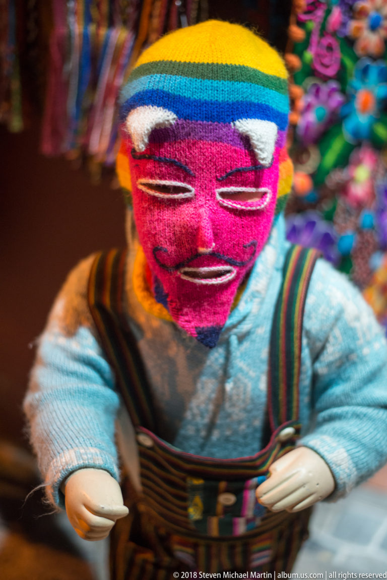 Colorful handmade doll with pink woven face, striped knitted hat, stitched features, and textile clothing.