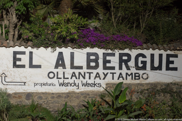 Painted wall sign reading “El Albergue Ollantaytambo,” with proprietor name, arrow, and flowers above.