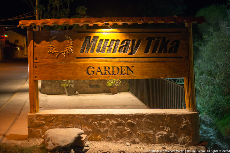 Wooden nighttime sign reading “Munay Tika Garden” above a stone base beside an entrance path.
