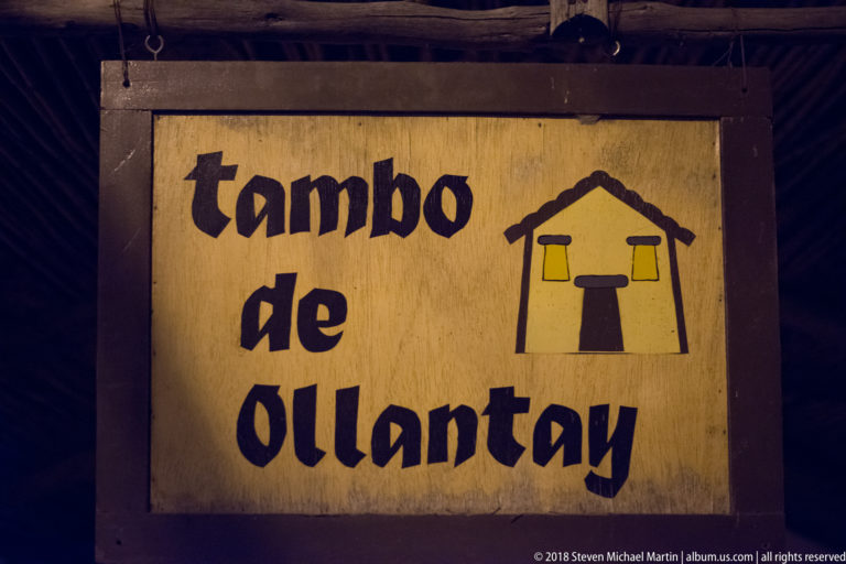 Wooden illuminated sign reading “tambo de Ollantay” beside a stylized yellow house emblem.