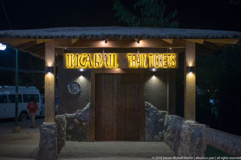 Night view of a small Inca Rail train ticket office with illuminated yellow signage above a wooden door.