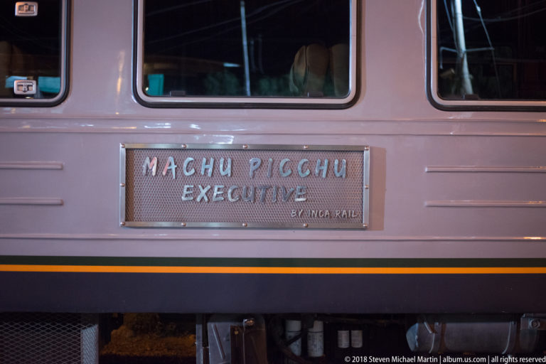 Metal train plaque reading “Machu Picchu Executive” and “by Inca Rail” on the side of a passenger coach.