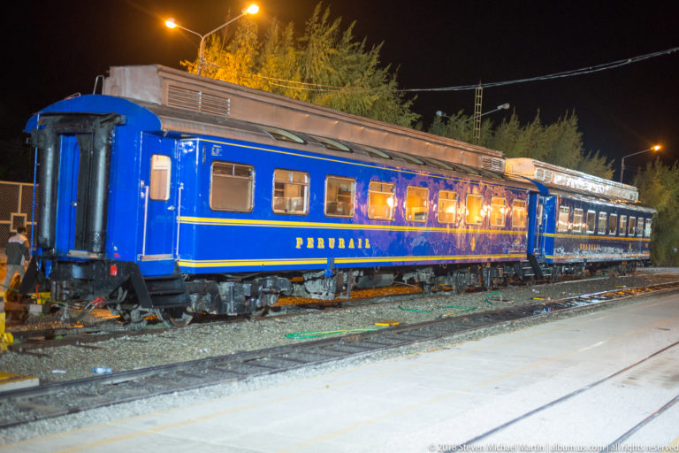 Blue PeruRail passenger train standing on tracks at night with illuminated windows and yellow trim.