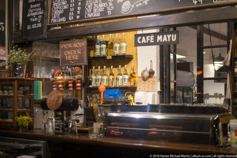 Café Mayu counter with espresso machine, bottles, glassware, hanging utensils, and chalkboard menu text.