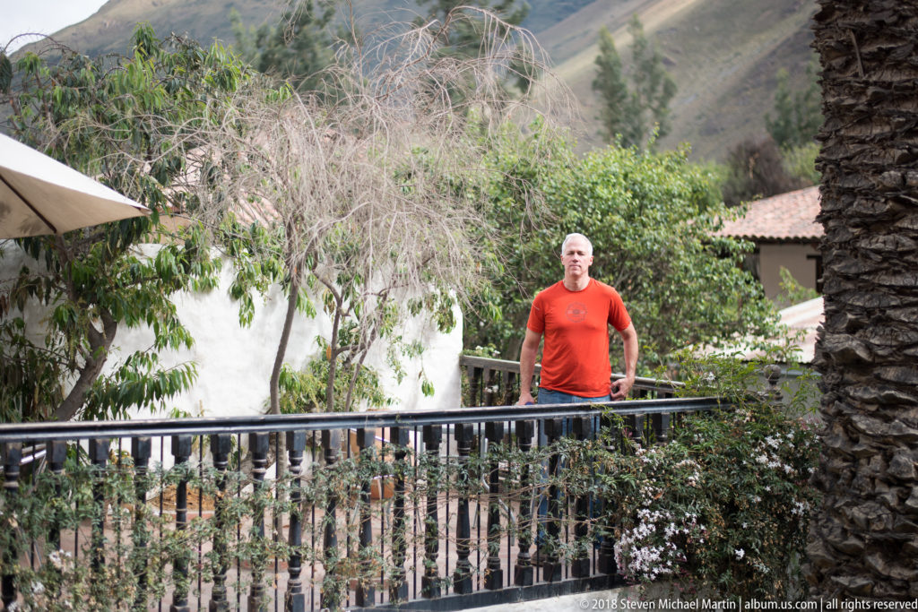 Man in a red shirt standing beside a terrace railing with trees, garden foliage, and mountain slope behind.