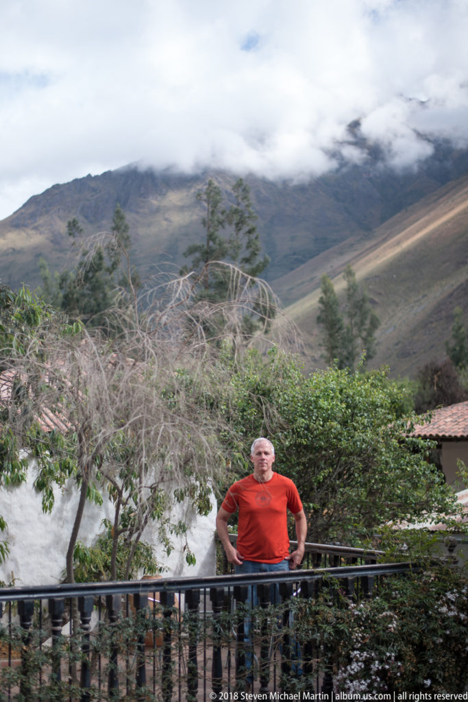 Man in a red shirt standing behind a railing with cloud-covered Andean mountains rising behind.