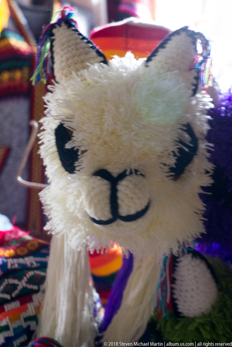 Close-up of a white plush alpaca toy with black stitched features amid colorful woven souvenirs.