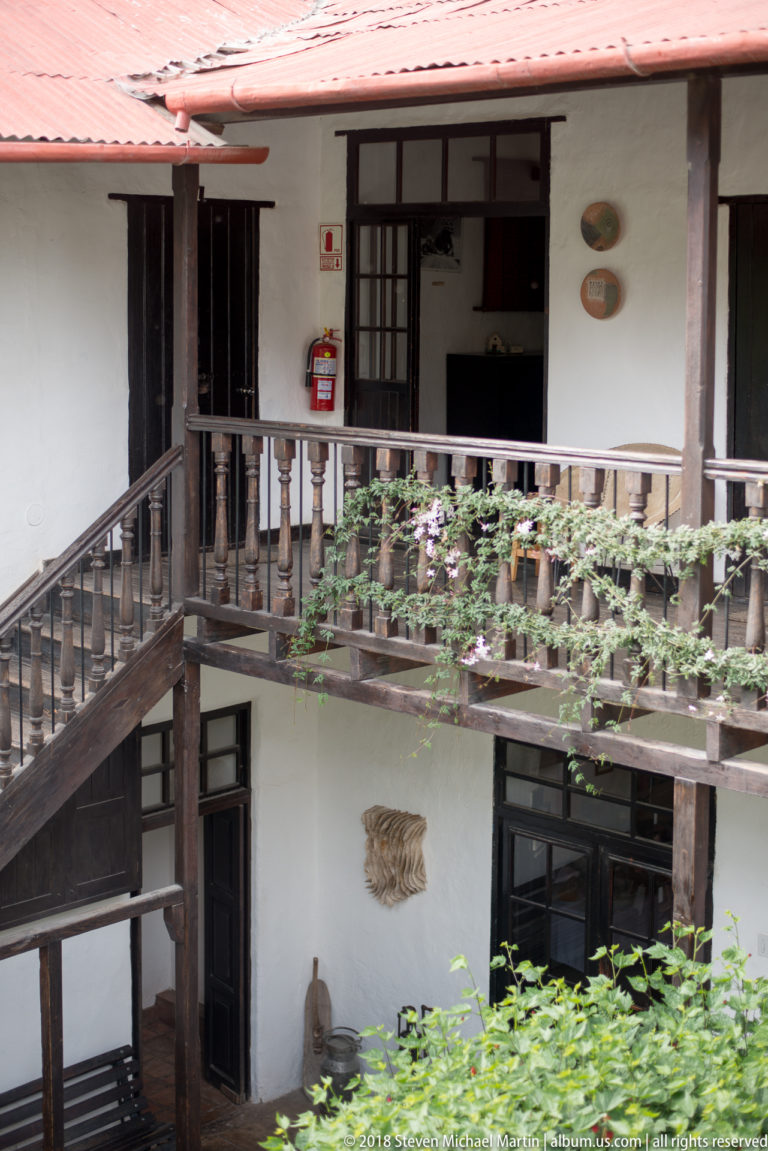 White-plastered courtyard with dark wooden balconies, staircase, doors, and rows of potted plants.