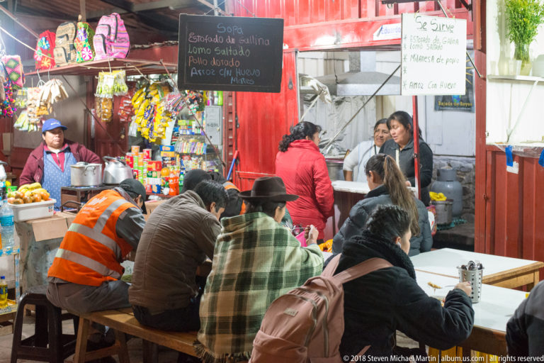 Customers seated at a small local eatery with red walls, counter service, snacks, fruit, and handwritten menu boards.