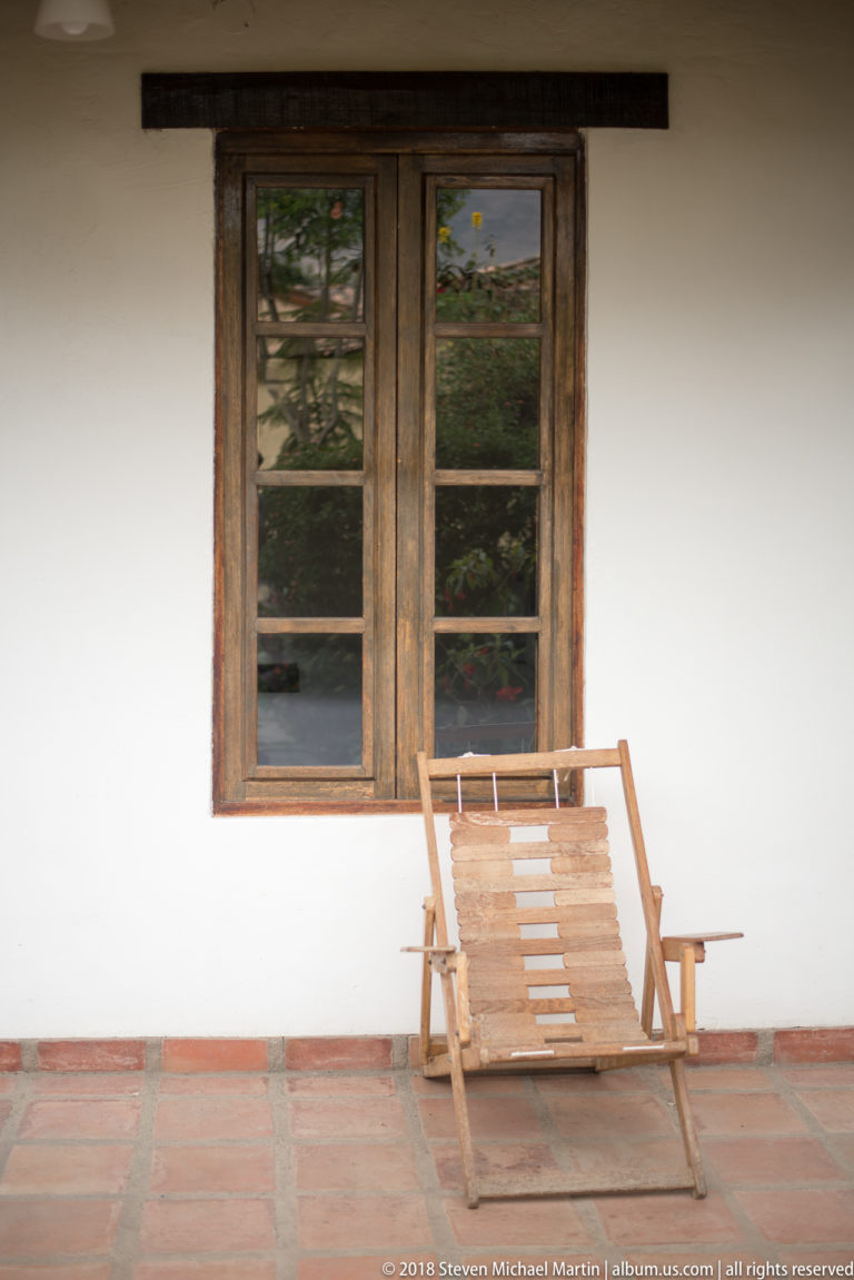 Wooden reclining chair on a terracotta-tiled veranda beneath a dark multi-pane window in a white wall.
