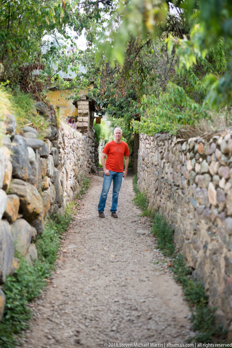 Man in a red shirt standing farther down a long path bordered by stone walls and overhanging foliage.