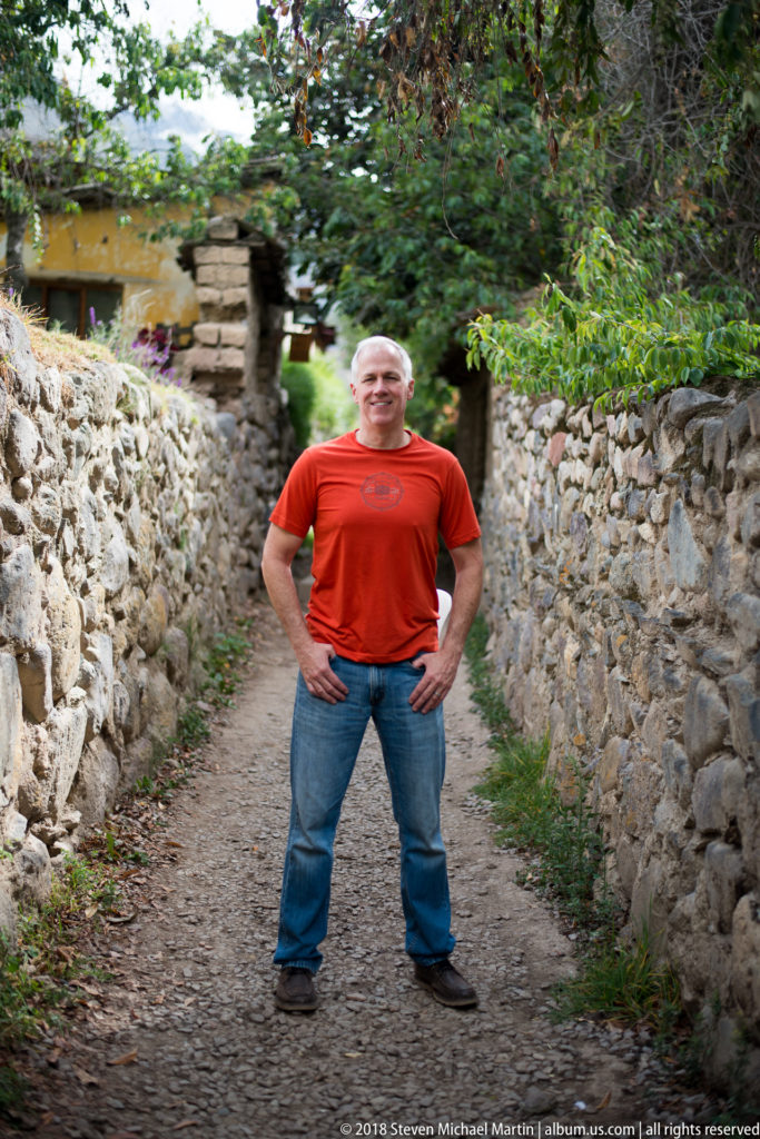 Man in a red shirt standing on a narrow path between rough stone walls beneath trees.