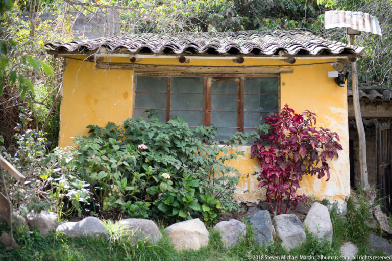 Small yellow plaster building with tile roof and dark windows surrounded by shrubs and garden stones.