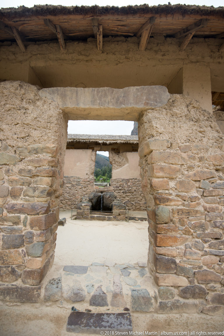 Aligned rectangular doorways through thick stone-and-earth walls, framing a distant fountain at Ollantaytambo.