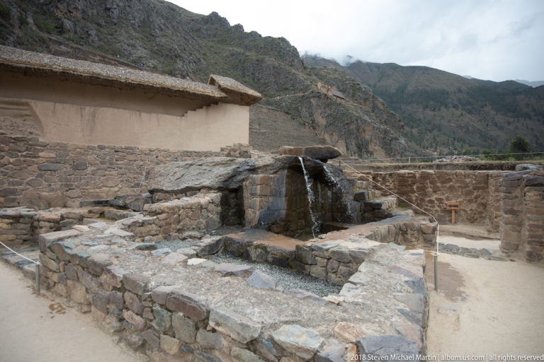 Water spilling over a stone fountain into a basin beside a roofed structure and low walls at Ollantaytambo.