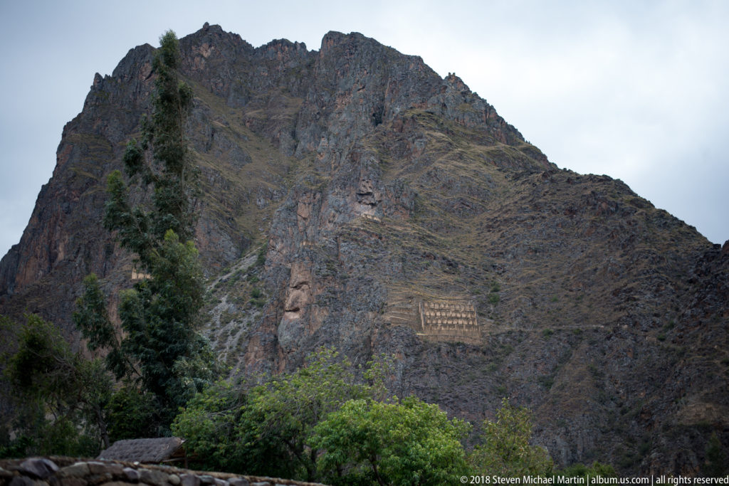Steep mountain face with long horizontal terraces and stone features above trees near Ollantaytambo.