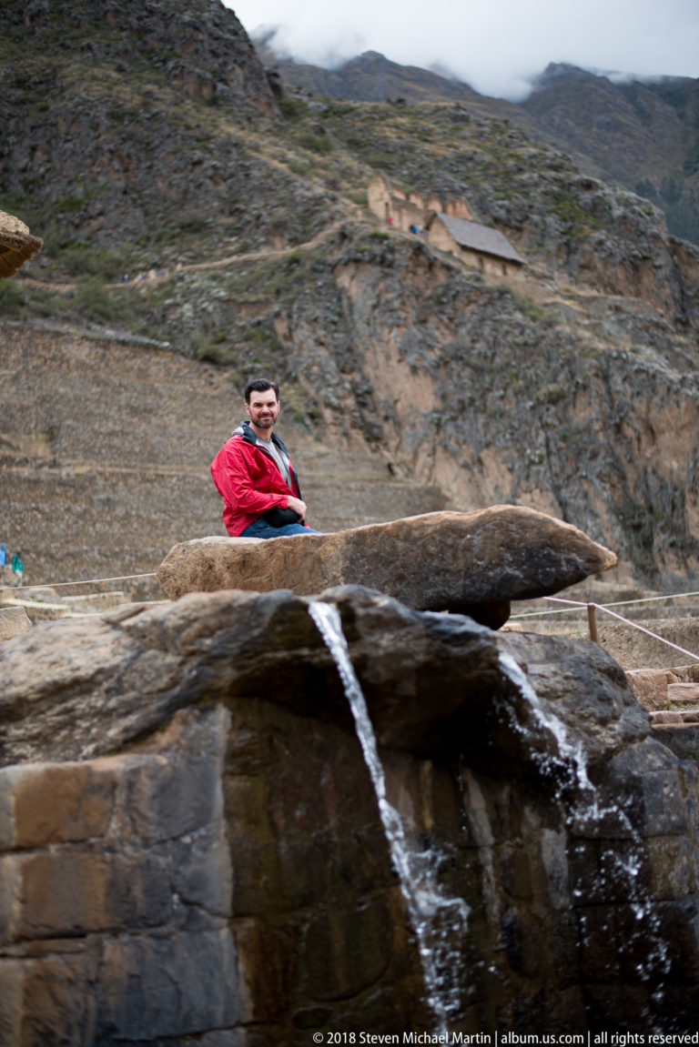Two streams of water spill beneath a long stone slab, with a visitor and terraces behind at Ollantaytambo.