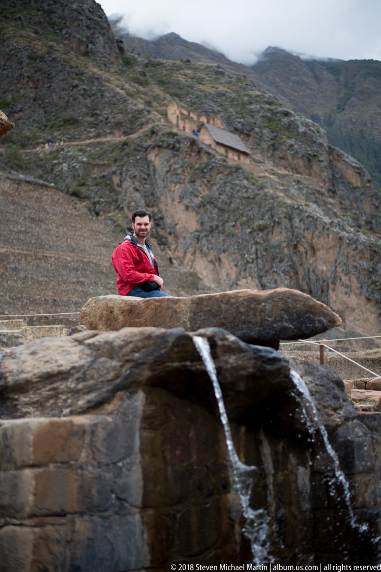 Water pouring from a stone outlet beneath a projecting slab, with a visitor standing behind at Ollantaytambo.
