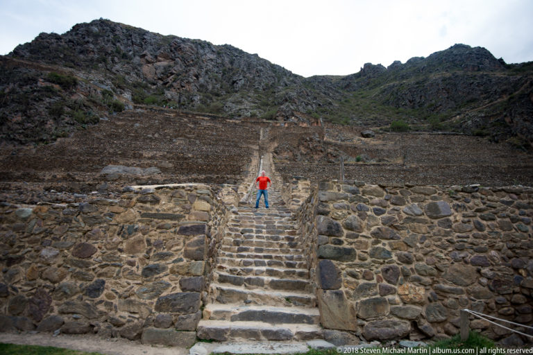 Stone staircase rising through multiple terrace walls at Ollantaytambo, with a visitor midway up.