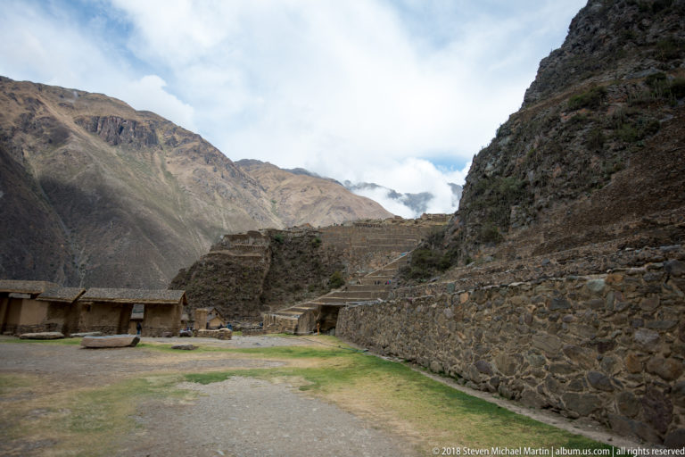 Open ground enclosed by stone walls and a thatched shelter beneath terraces at Ollantaytambo.