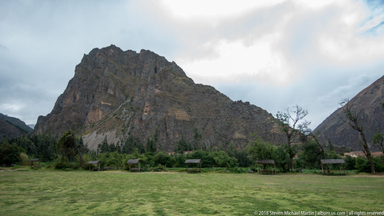 Steep mountain with visible terrace lines rising behind a grassy area and small shelters at Ollantaytambo.