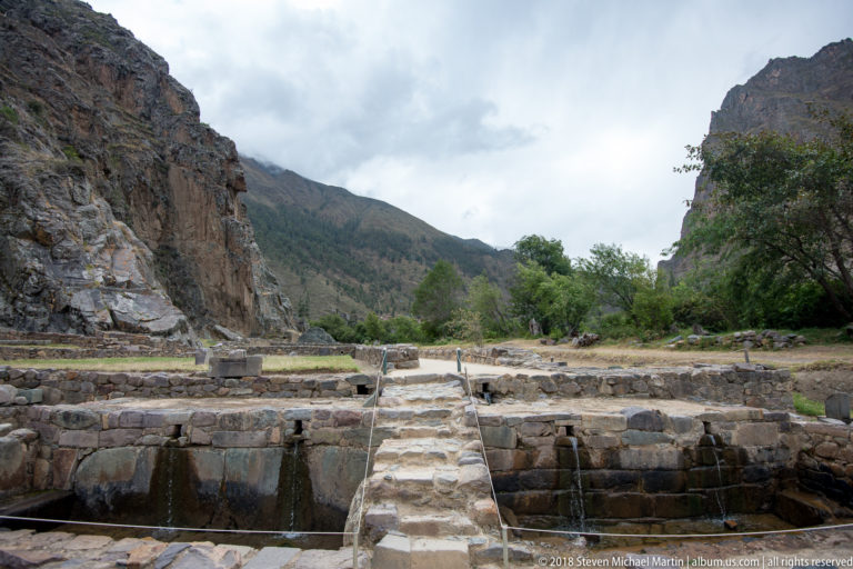 Rectangular stone-lined basins with flowing water and a narrow central crossing at Ollantaytambo.
