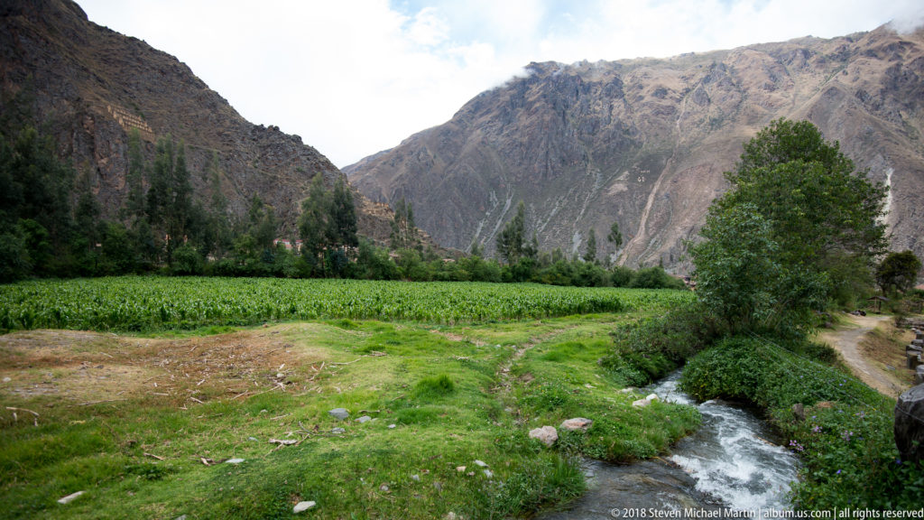 Green agricultural fields, a narrow stream, trees, and steep mountains in the Ollantaytambo valley.